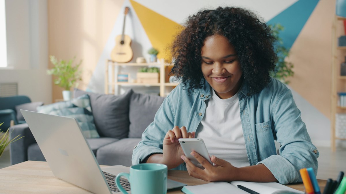 Estudante sorrindo enquanto usa o celular ao lado do notebook e do caderno